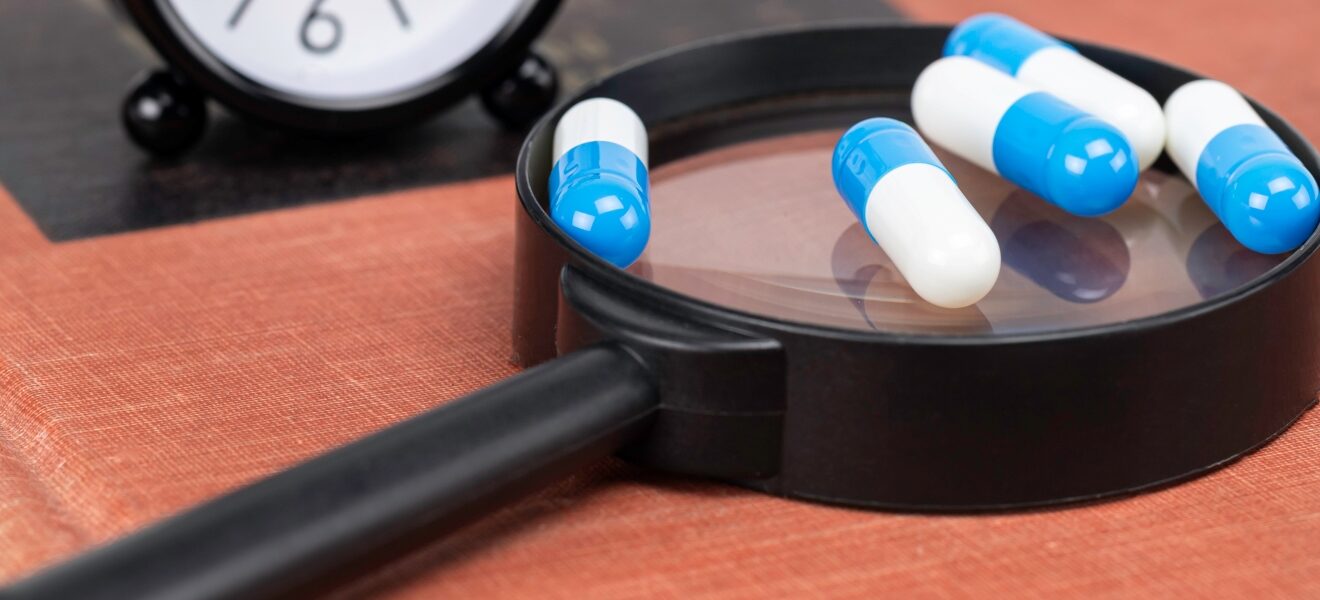 Four pills sitting on top of a magnifying glass with a clock in the background.
