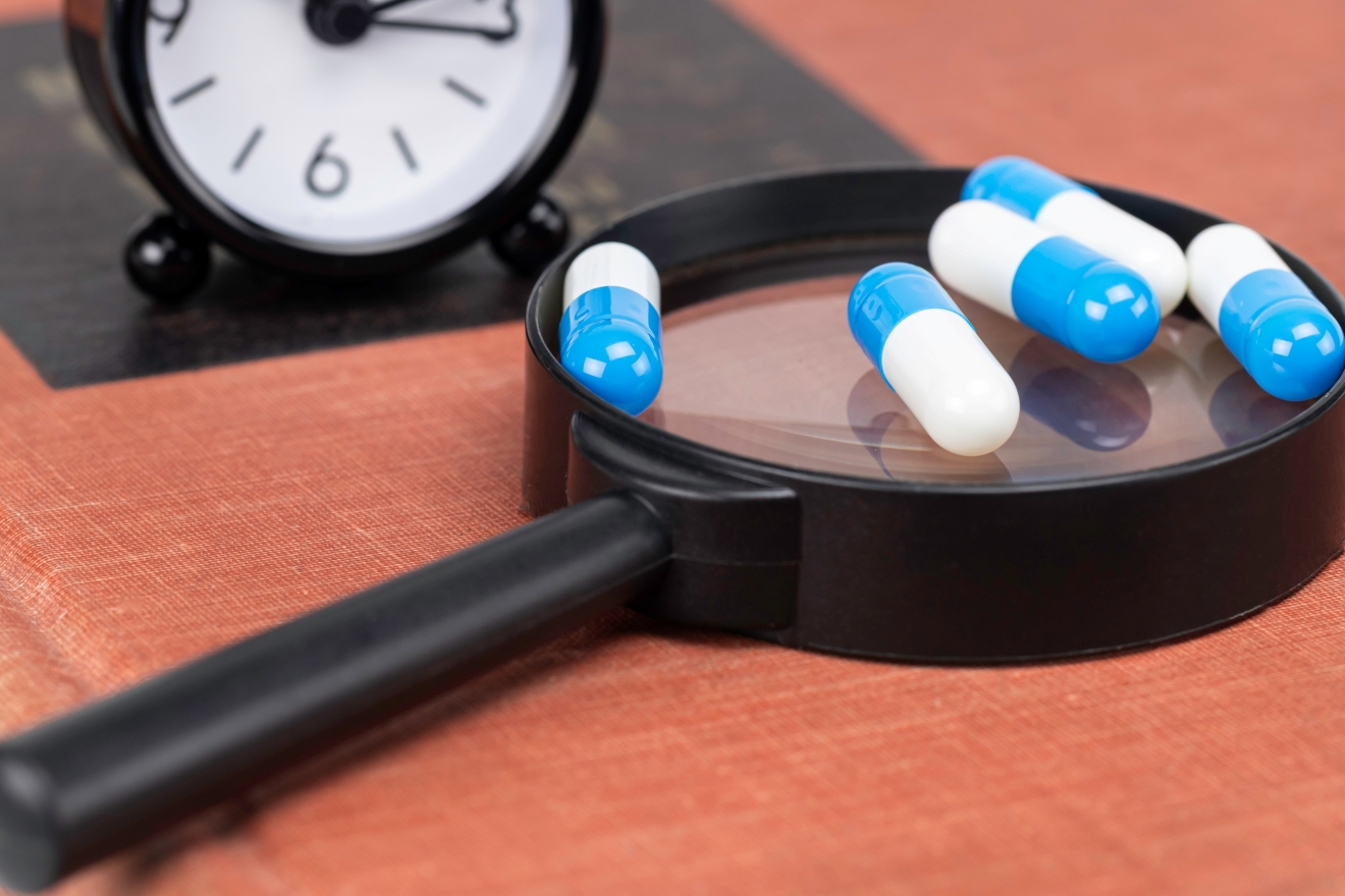 Four pills sitting on top of a magnifying glass with a clock in the background.