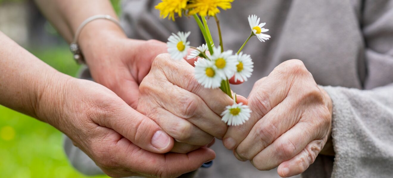 The hands of an older adult holding yellow and white flowers, their hands embraced by another adult.