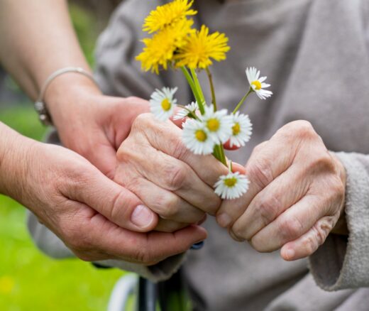 The hands of an older adult holding yellow and white flowers, their hands embraced by another adult.