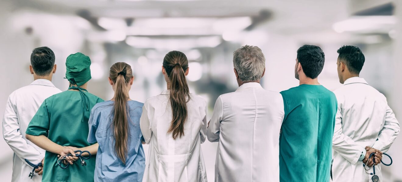 A team of clinicians in the ER standing shoulder-to-shoulder with their backs facing the camera.