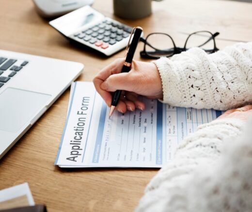 Person filling out application form on paper next to a laptop, calculator, and glasses on a table.