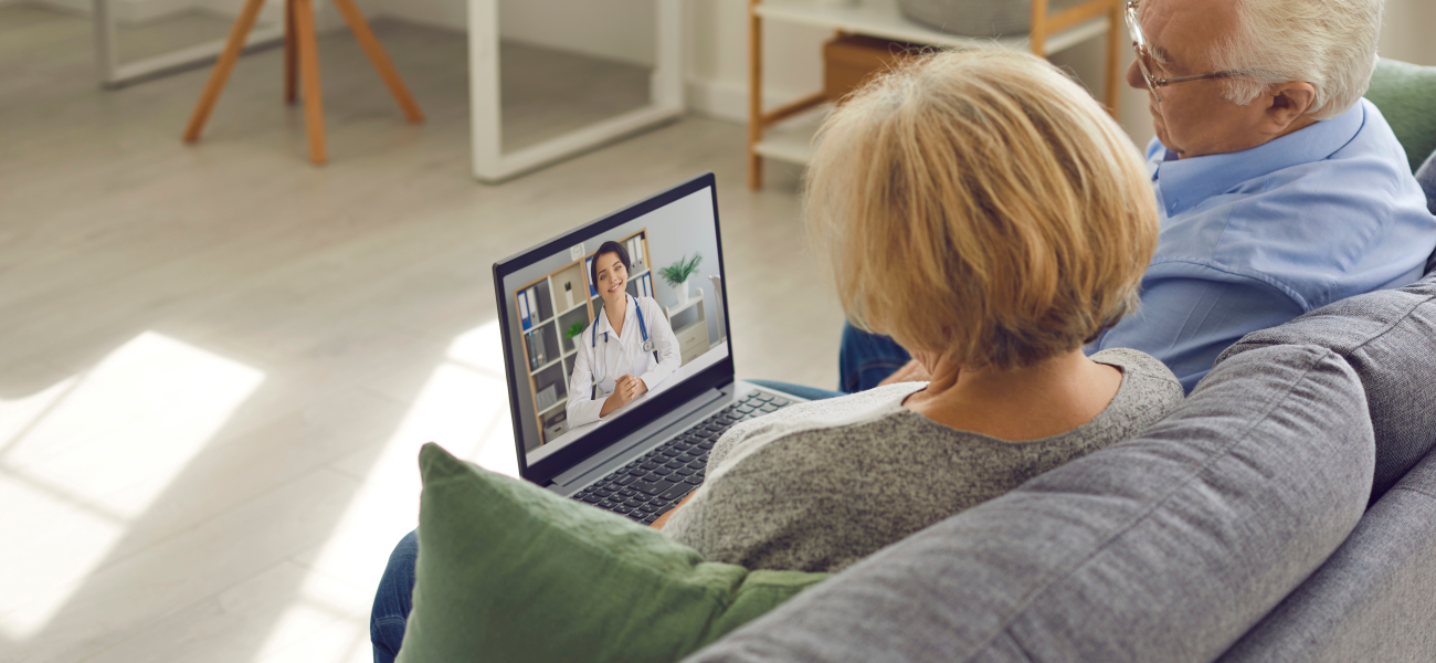 An elder man and woman sit on the couch, attending a telehealth appointment on a laptop.