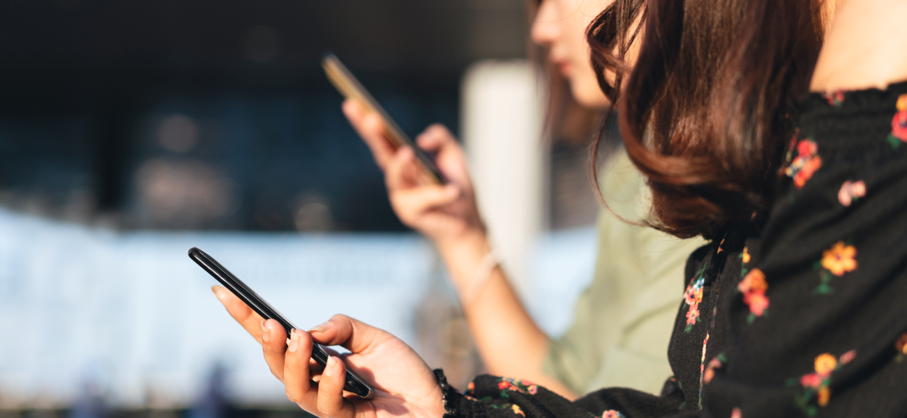 Two women sitting side by side, one in the background, both on their mobile devices.