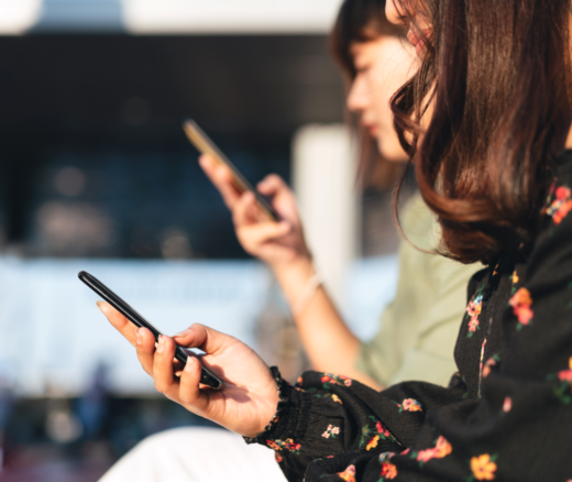 Two women sitting side by side, one in the background, both on their mobile devices.