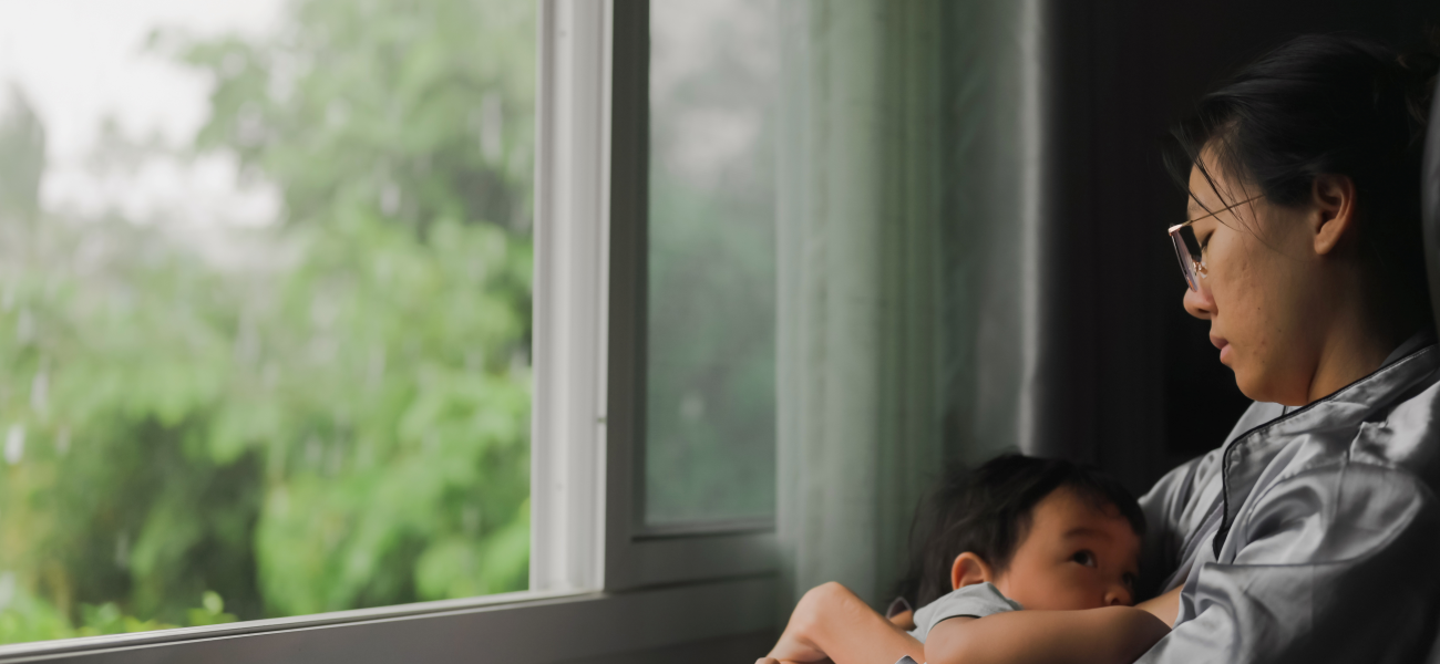 An Asian woman sitting in a chair facing a window with her infant sitting in her lap.