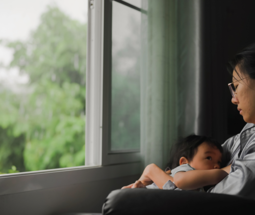 An Asian woman sitting in a chair facing a window with her infant sitting in her lap.