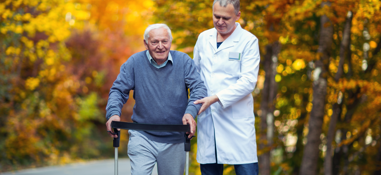 An older adult with a walker being led by a caretaker on a street with fall foliage.