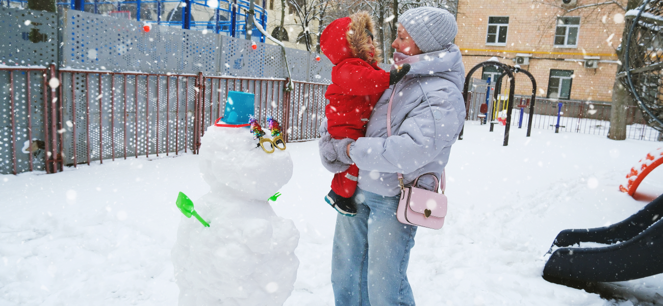 An infant and their mother bundled up for snowy weather at a park next to a snowman.