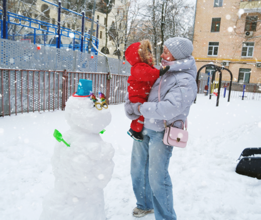 An infant and their mother bundled up for snowy weather at a park next to a snowman.