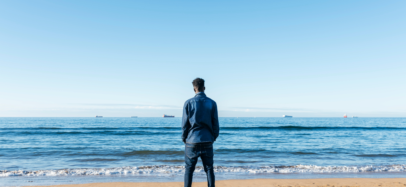 An African man on the beach looking at the ocean from behind.